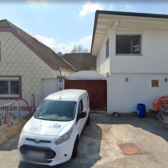 Ein weißer Ford-Transporter ist vor einem Haus in einer Einfahrt geparkt. Das Haus hat ein braune Tor und Fenster. In der Nähe befinden sich Fässer und ein Schubkarren.