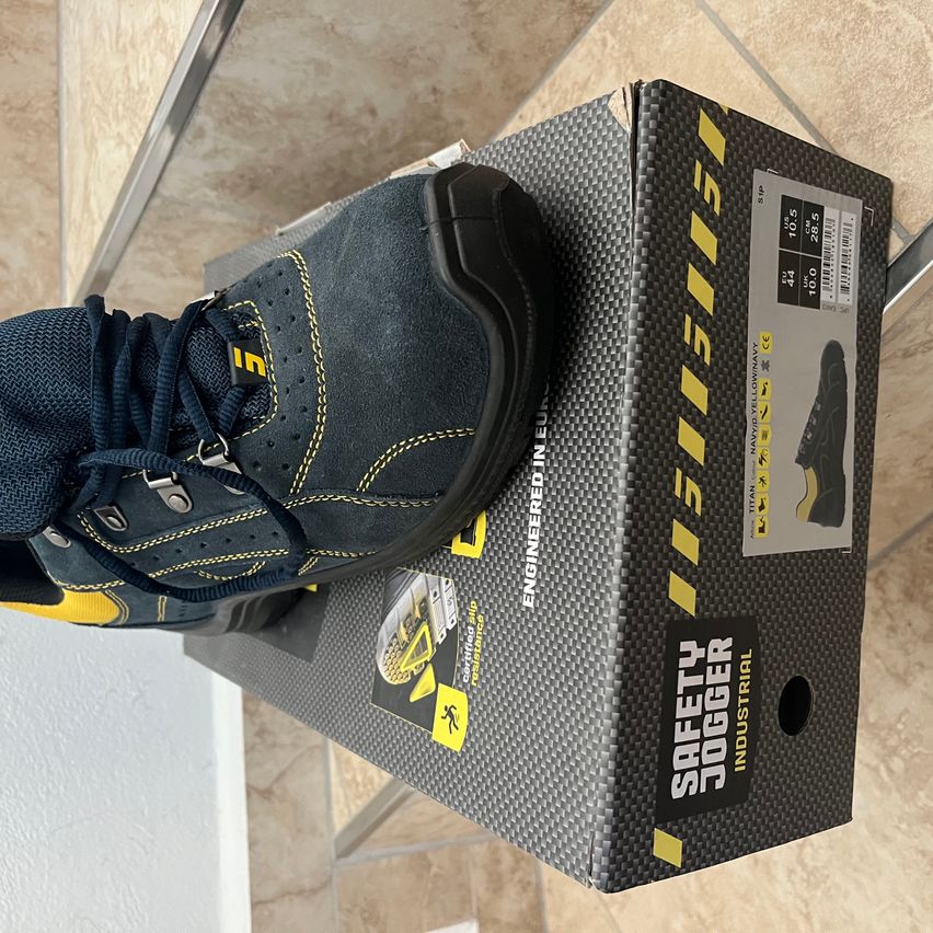 Ein Paar navyblaue und gelbe Sicherheitsschuhe mit schwarzer Sohle liegen auf einem schwarzen Schuhkarton mit gelbem und schwarzem Design.