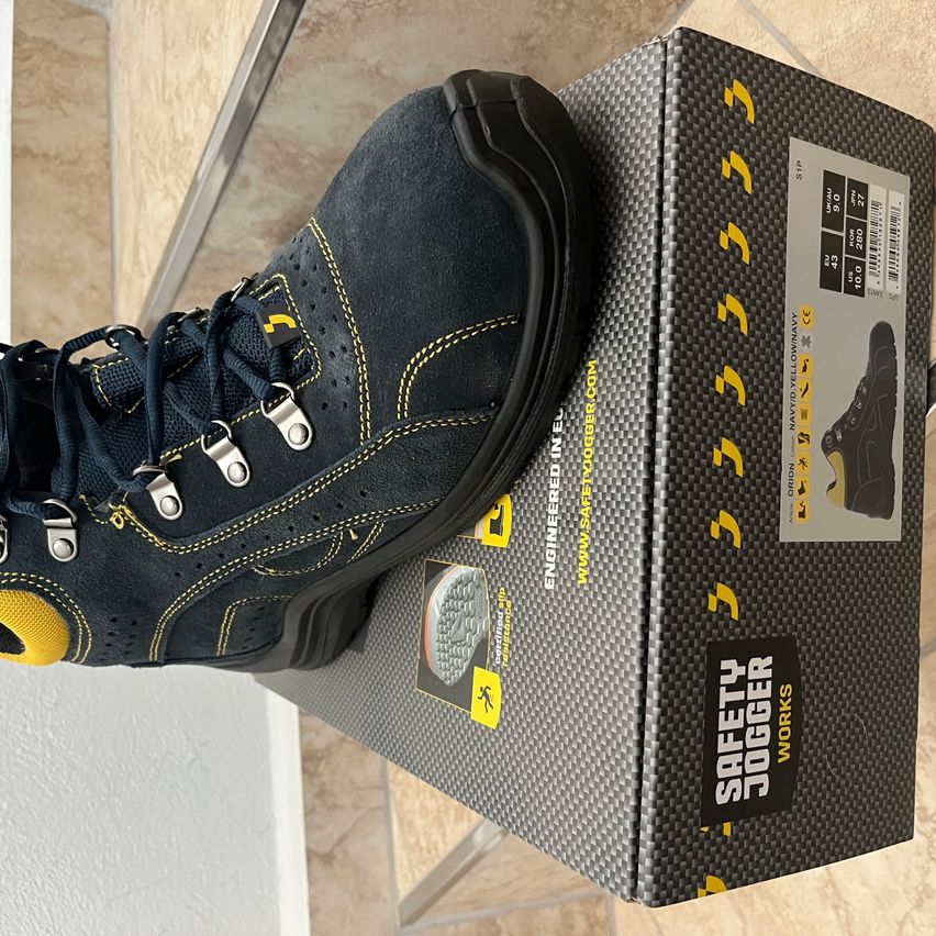 Ein Paar navyblaue und gelbe Arbeitsschuhe mit Metallschnallen liegt auf seiner Schachtel. Die Schachtel hat ein schwarzes und gelbes Design mit dem Markennamen Safety Jogger.