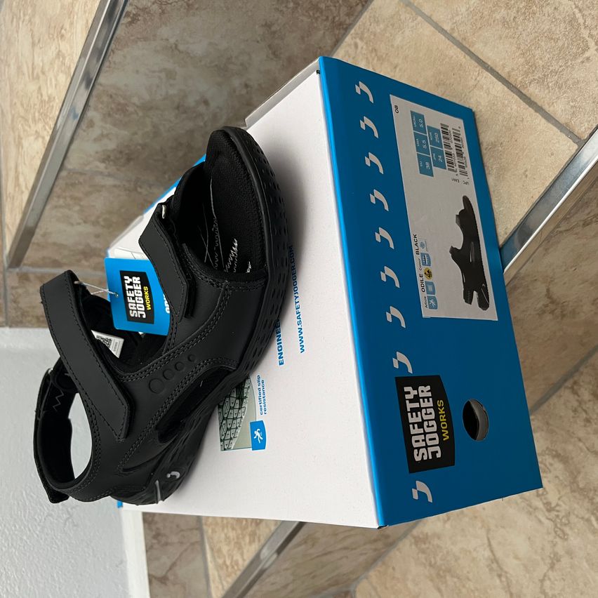 Ein schwarzer Sandalen liegt auf seiner Schachtel, die den Text 'Safety Jogger' und 'ENGINEERED FOR WORK' aufweist.