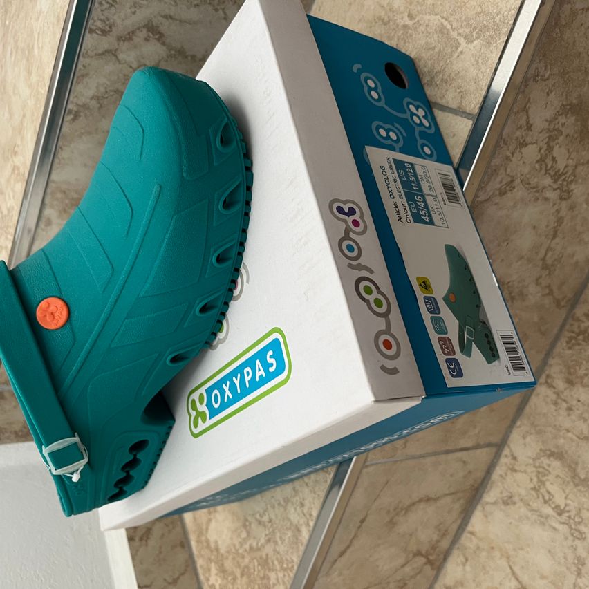 Ein türkisfarbener Oxypas-Schuh liegt auf seiner Verpackung, die das Markenlogo und Produktdetails zeigt. Der Schuh hat eine strukturierte Sohle und einen verstellbaren Riemen.