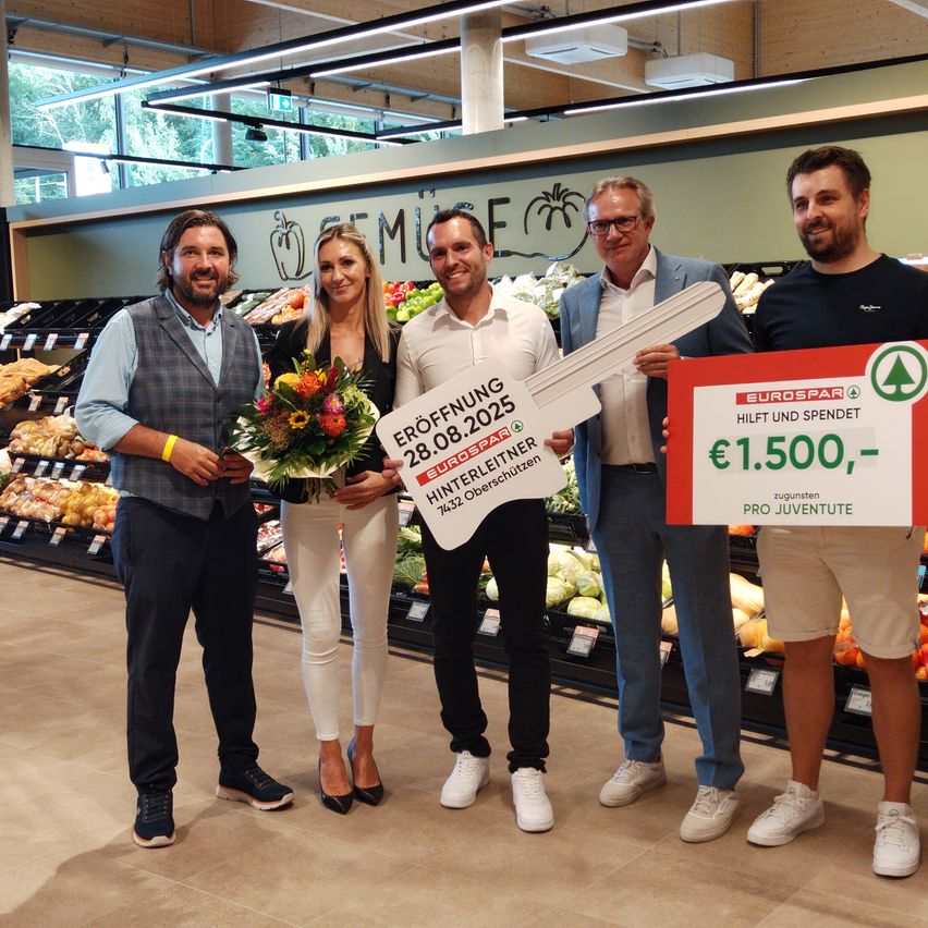 Fünf Personen stehen in einem Lebensmittelgeschäft, halten einen Blumenstrauß und einen Schlüssel mit Text darauf. Die Person ganz rechts hält ein rotes Schild, das 'Hilft und Spendet' sagt.