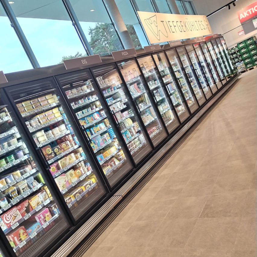 Ein Supermarktregal mit gekühltem Sortiment an Lebensmitteln, beschriftet mit 'EIS', 'PIZZA' und 'TEEFGEKÜHLTES'. Darüber befindet sich ein Schild mit Kuh-Symbol.