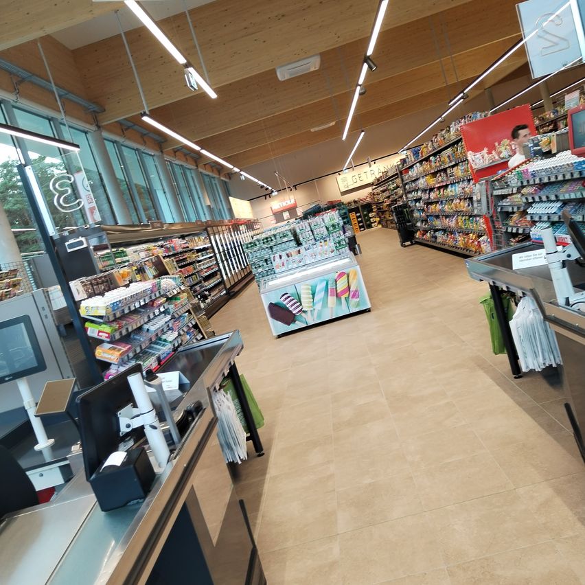 Ein leerer Supermarktgang mit einer Vielzahl von Produkten in den Regalen. Eine Person steht neben einem roten Regal, und eine Ausstellung von Eis am Stiel ist vorhanden.
