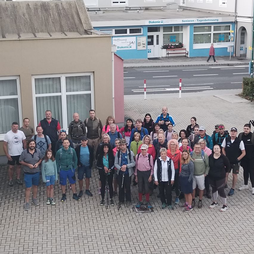 Eine Gruppe von Menschen ist vor einem Gebäude mit einer blau-weißen Wand versammelt, wahrscheinlich für eine Gemeinschaftsveranstaltung. Dahinter ist eine Straße zu sehen, auf der eine Person auf dem Bürgersteig geht.