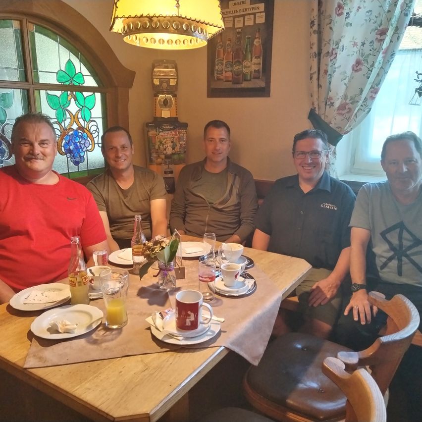 Fünf Männer sitzen lächelnd um einen Tisch herum, die Kamera im Blick. Der Tisch ist mit Tellern, Tassen und Flaschen gedeckt. Das Zimmer hat ein Fenster mit einem Blumenvorhang und eine Lampe, die von der Decke hängt. Ein gerahmter Werbeplakat ist an der Wand angebracht.