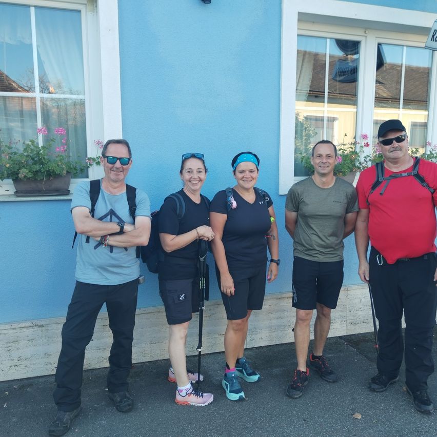 Fünf Personen, wahrscheinlich Wanderer, stehen vor einem blauen Gebäude mit Fenstern und lächeln für ein Foto. Sie tragen alle Rucksäcke und Sportschuhe.