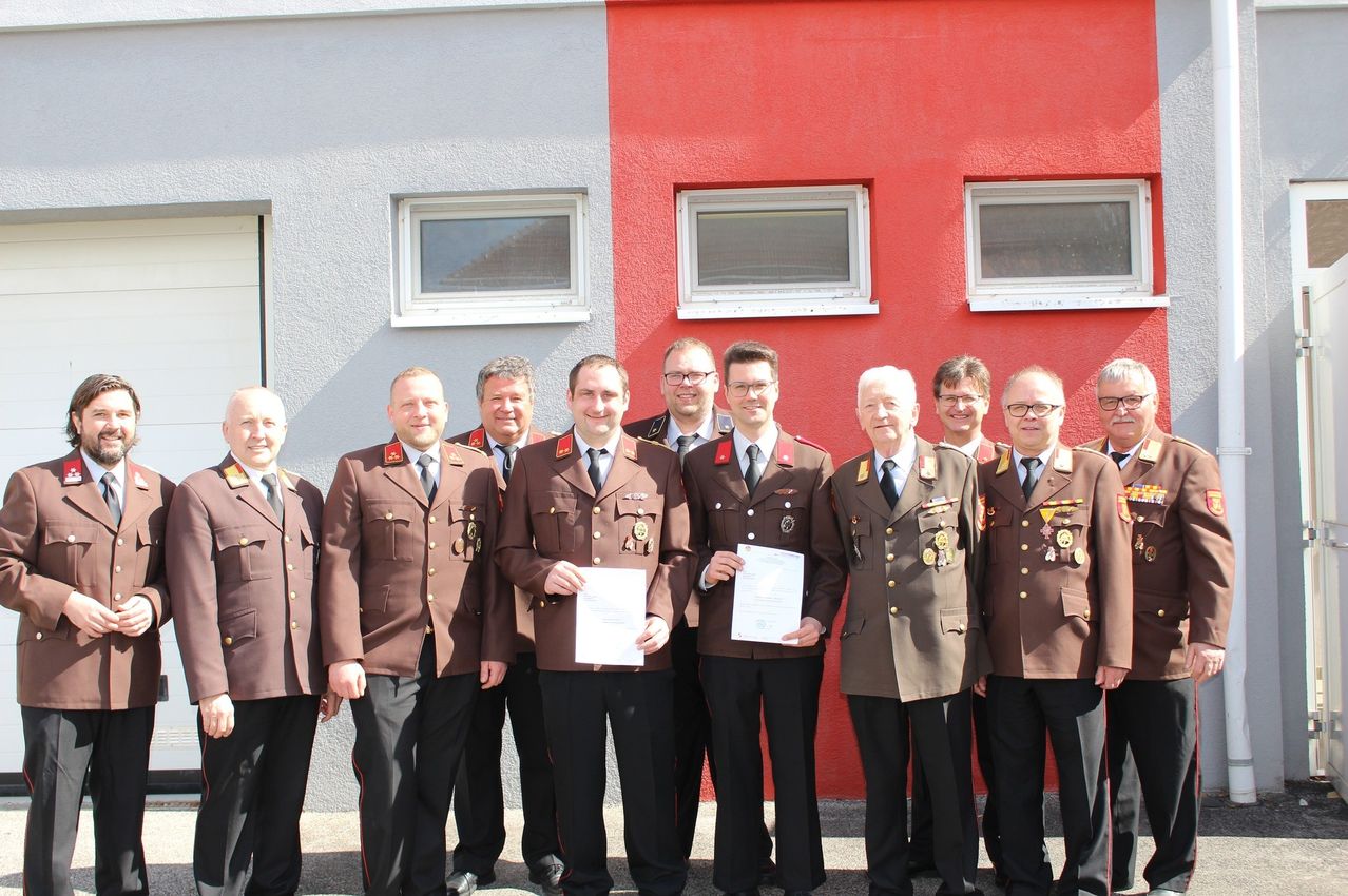 Eine Gruppe von Männern in Militäruniformen steht vor einem roten Gebäude mit drei Fenstern. Sie halten Papiere in den Händen und lächeln.