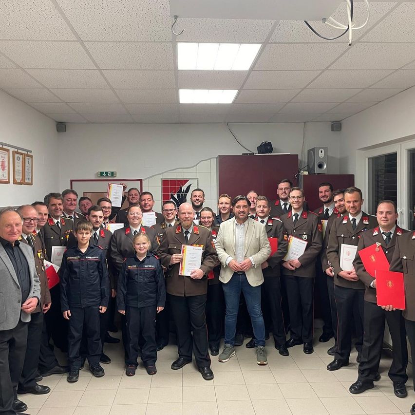 Eine Gruppe von Militärangehörigen und Beamten steht zusammen in einem Raum und hält Urkunden. Ein Mann in einem weißen Blazer steht zentral und lächelt. Dahinter stehen in Reihen Uniformierte.