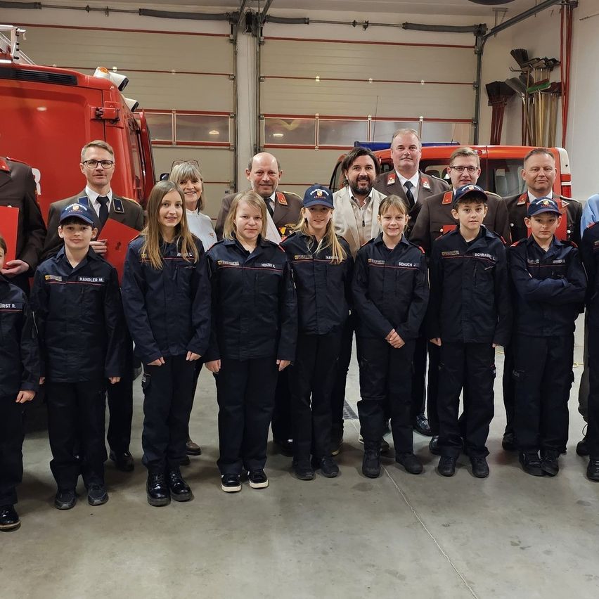 Eine Gruppe von Kindern und Erwachsenen in Uniform posiert für ein Foto in einer Feuerwache, mit einem Feuerwehrauto im Hintergrund.