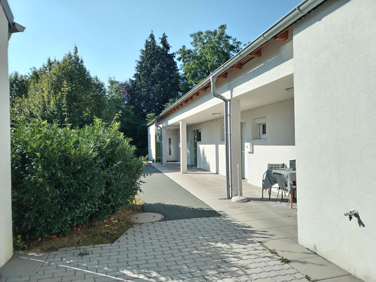 Ein modernes weißes Gebäude mit einem gepflasterten Weg, Bäumen und einem Tisch mit einem Stuhl draußen.