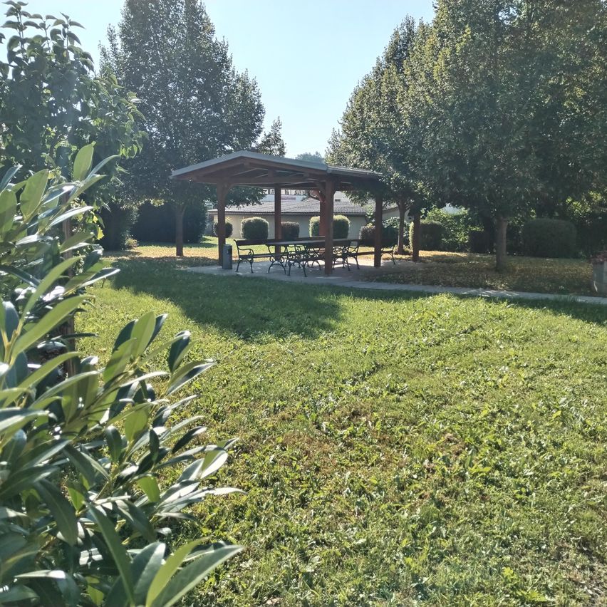 Ein hölzerner Pavillon steht inmitten eines grünen Rasens mit einem Tisch und Stühlen. Der Pavillon ist von Büschen und Bäumen umgeben. Die Sonne scheint hell, und der Himmel ist klar. In der Ferne befindet sich ein Haus mit einem grauen Dach.