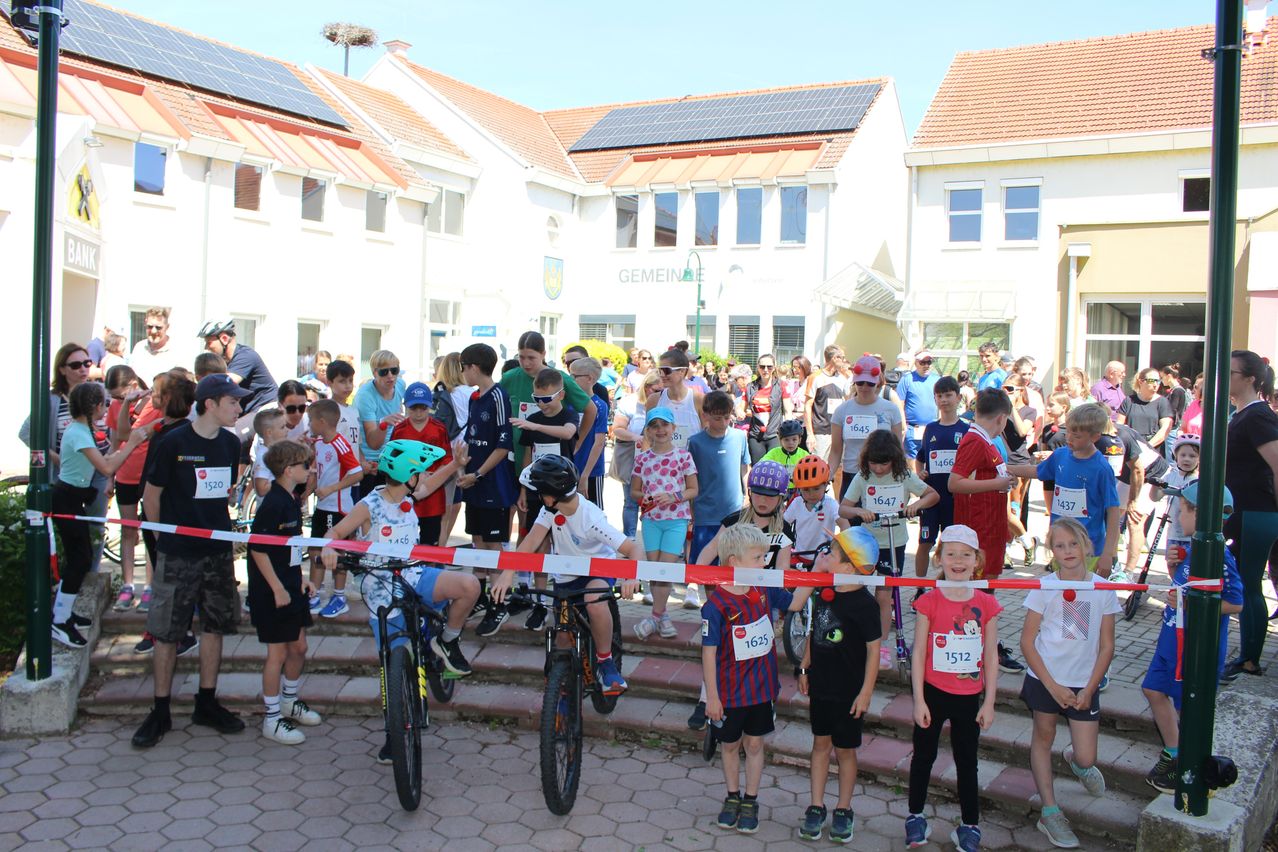 Eine Gruppe von Kindern in Helmen wartet in einer Schlange für ein Fahrradrennen. Die Kinder stehen vor Gebäuden mit Solarpanelen auf den Dächern.