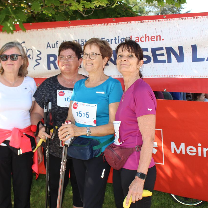 Vier Frauen stehen vor einem Banner für einen Marathon. Sie tragen alle eine Brille und halten Stöcke und eine Banane. Die Frau auf der linken Seite hat ein rotes Band.