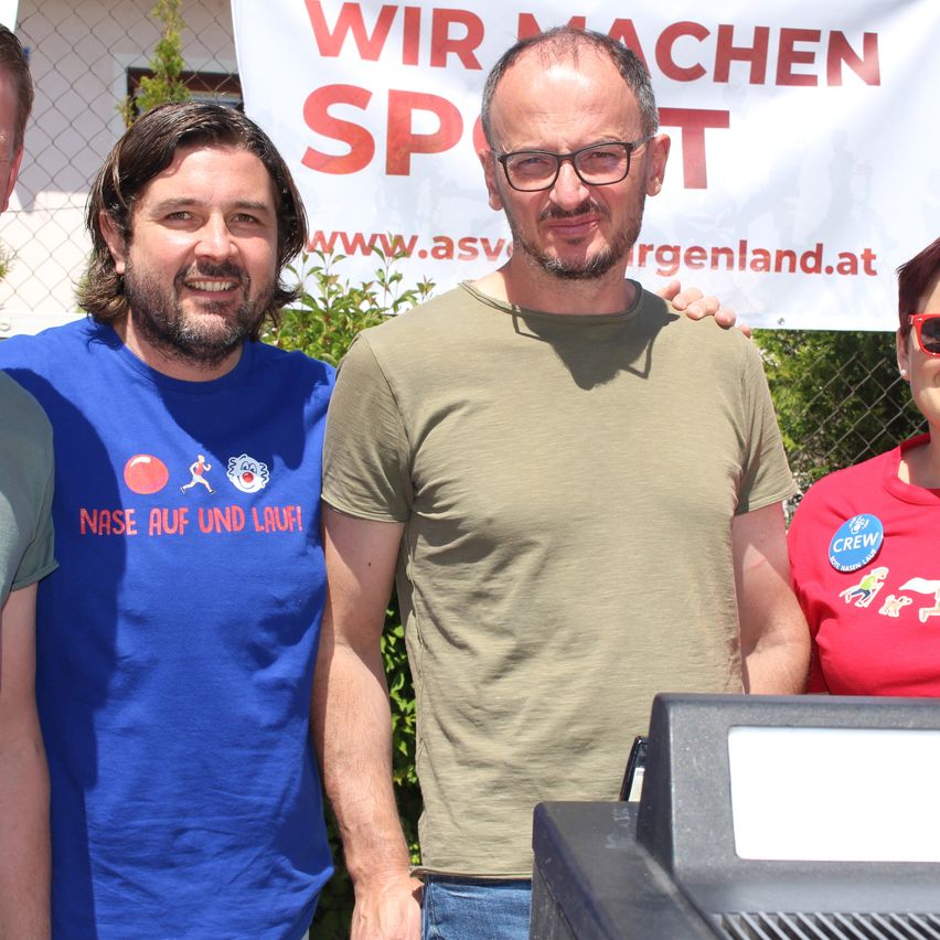Drei Männer stehen lächelnd vor einem Zaun. Einer trägt ein blaues Shirt mit 'Nase auf und Lauf!' Der Mann in der Mitte hat eine Brille und ein grünes Shirt. Eine Frau in einem roten Shirt steht rechts. Hinter ihnen ist ein Banner mit 'Wir machen Sport'.