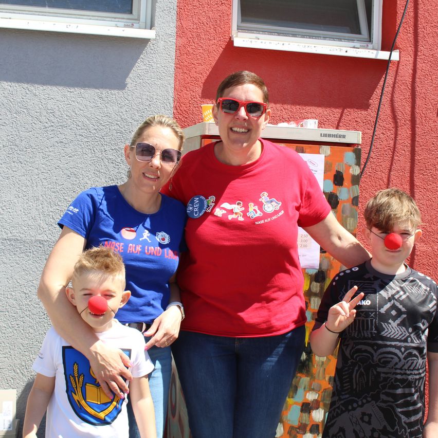 Eine Frau in einem blauen Shirt und zwei Kinder mit Clownsnasen stehen vor einem roten Gebäude. Eine weitere Frau in Rot lächelt und posiert mit den Armen um die Kinder.