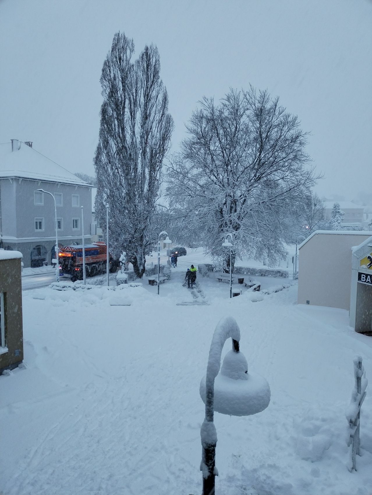 Eine verschneite Straßenszene mit einem roten Schneepflug, Menschen in gelben Westen und schneebedeckten Bäumen. Straßenlaternen und Bänke sind ebenfalls mit Schnee bedeckt.