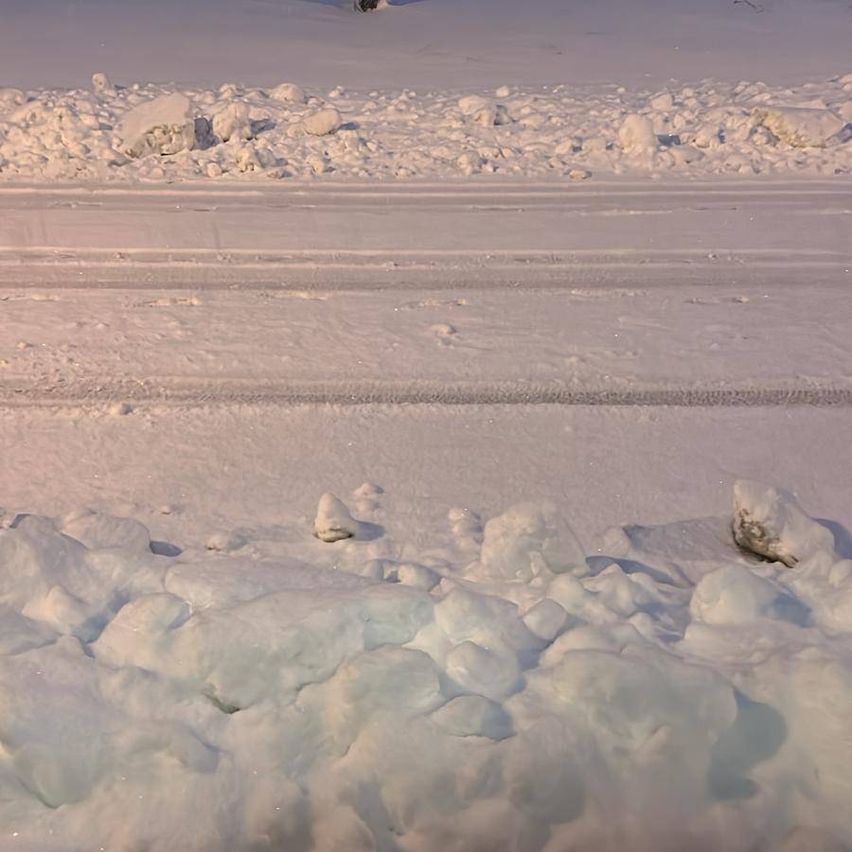 Eine verschneite Landschaft mit einer Straße in der Mitte, bedeckt mit Schnee und Eis. Schneehaufen sind auf beiden Seiten der Straße verstreut.