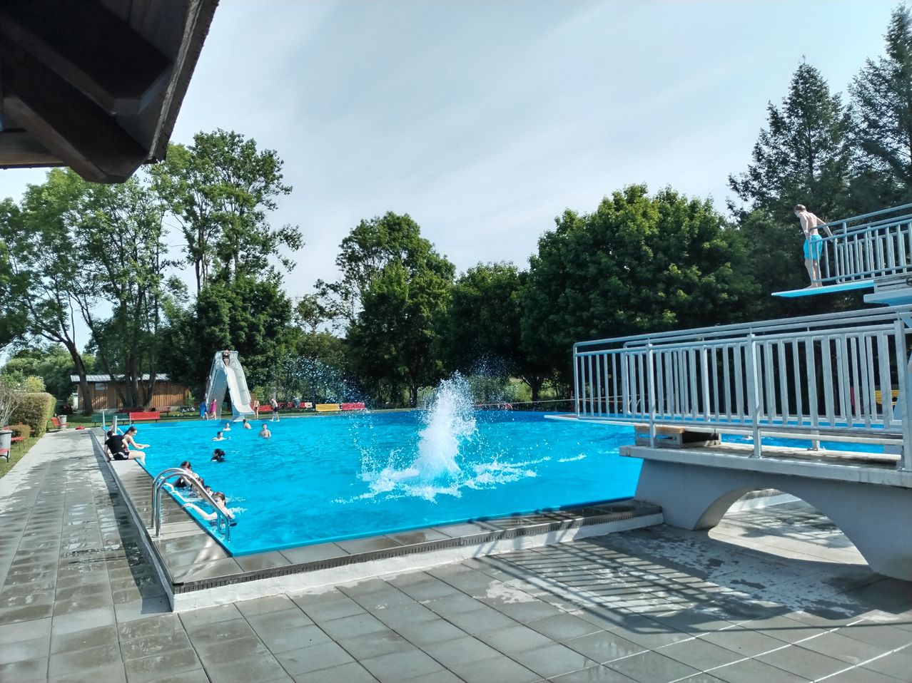 Ein Freibad mit schwimmenden Menschen und einem Wasserspritzer. Rechts befinden sich eine Rutsche und ein Sprungbrett. Bäume umgeben das Gelände.