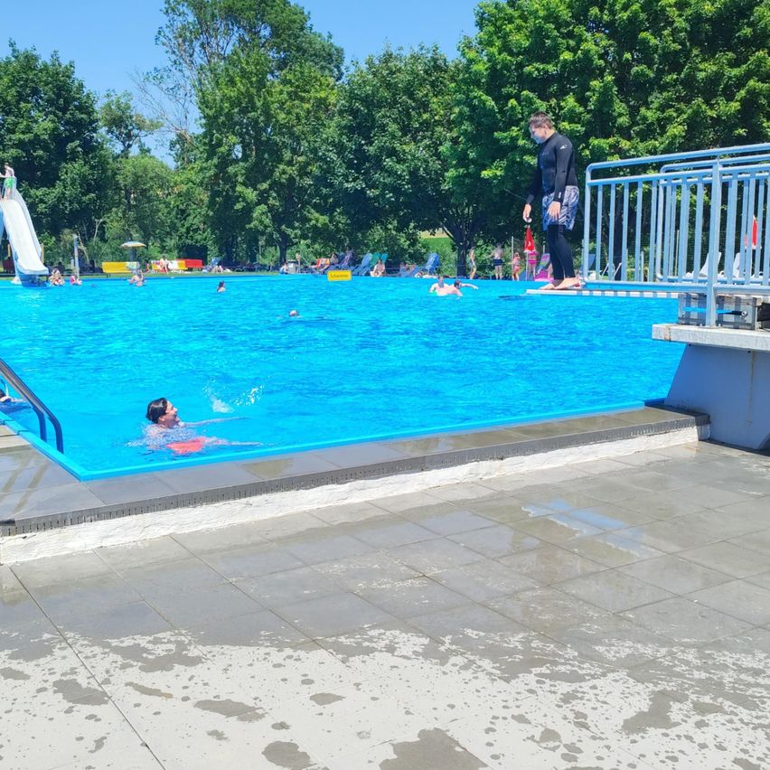 Ein großer Swimmingpool mit einem Mann, der an der Seite steht. Viele Menschen schwimmen im Pool und einige sind auf der Rutsche. Bäume und Gras sind im Hintergrund.