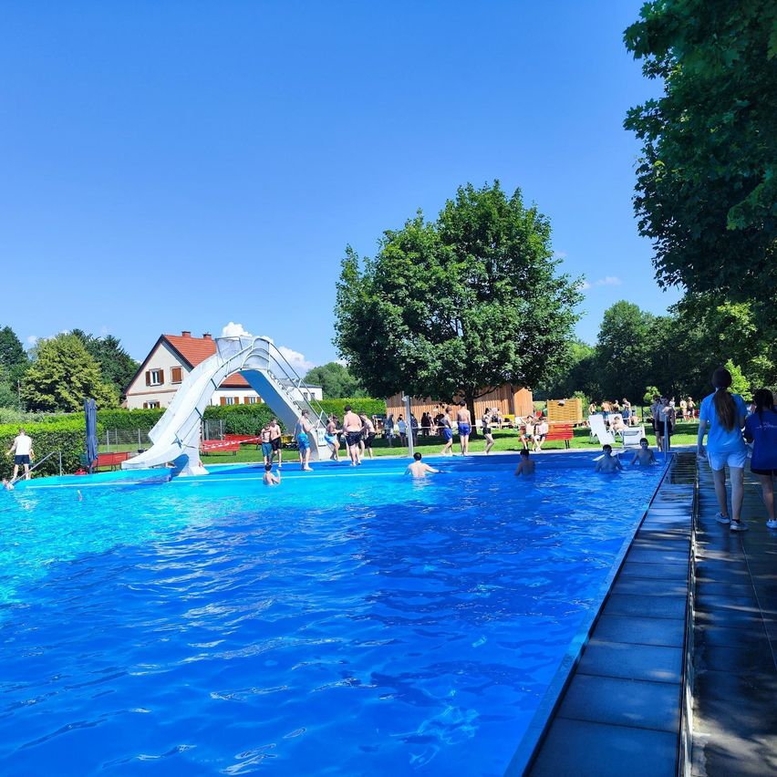 Ein Freibad voller Menschen. Einige schwimmen, andere gehen herum. Eine weiße Rutsche befindet sich an der Seite.