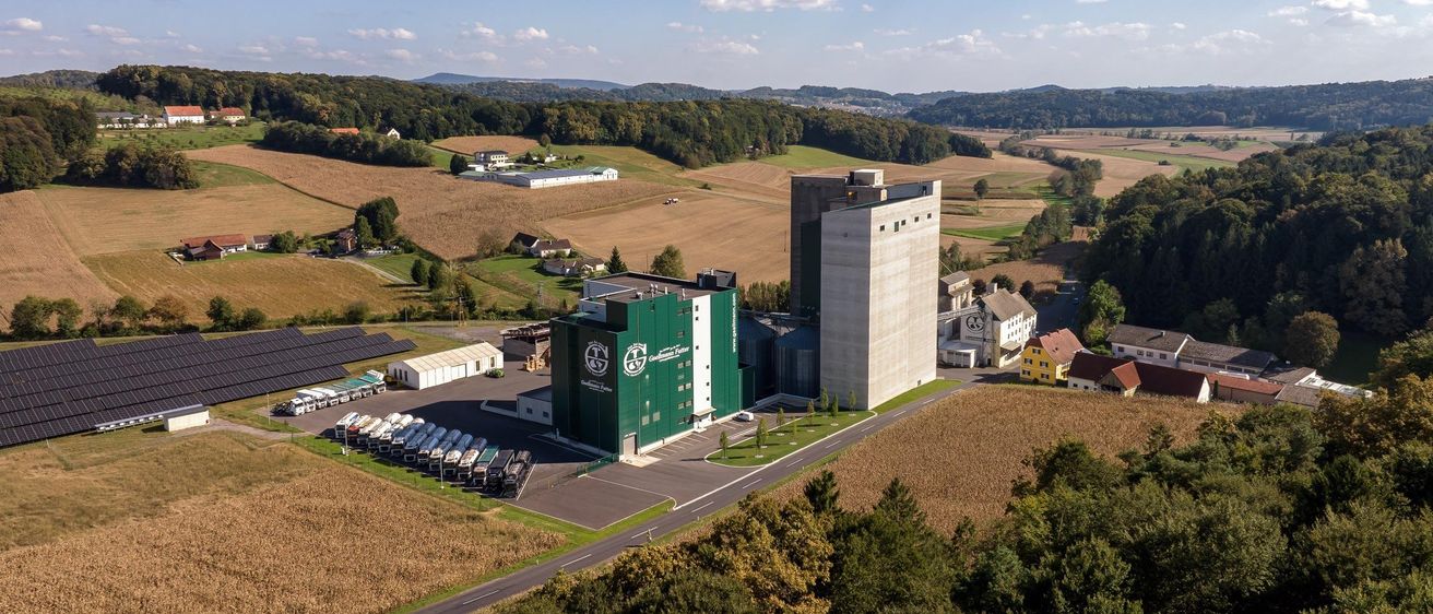 Luftaufnahme einer Industrieanlage mit einem großen grünen Gebäude und einem hohen Silo. Das Gebiet ist von Feldern, Bäumen und Häusern umgeben.