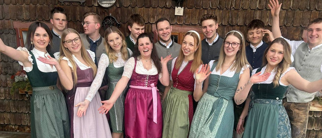 Eine Gruppe von Menschen in traditioneller deutscher Kleidung, die lächelnd und posierend für ein Foto vor einem Holzgebäude stehen.
