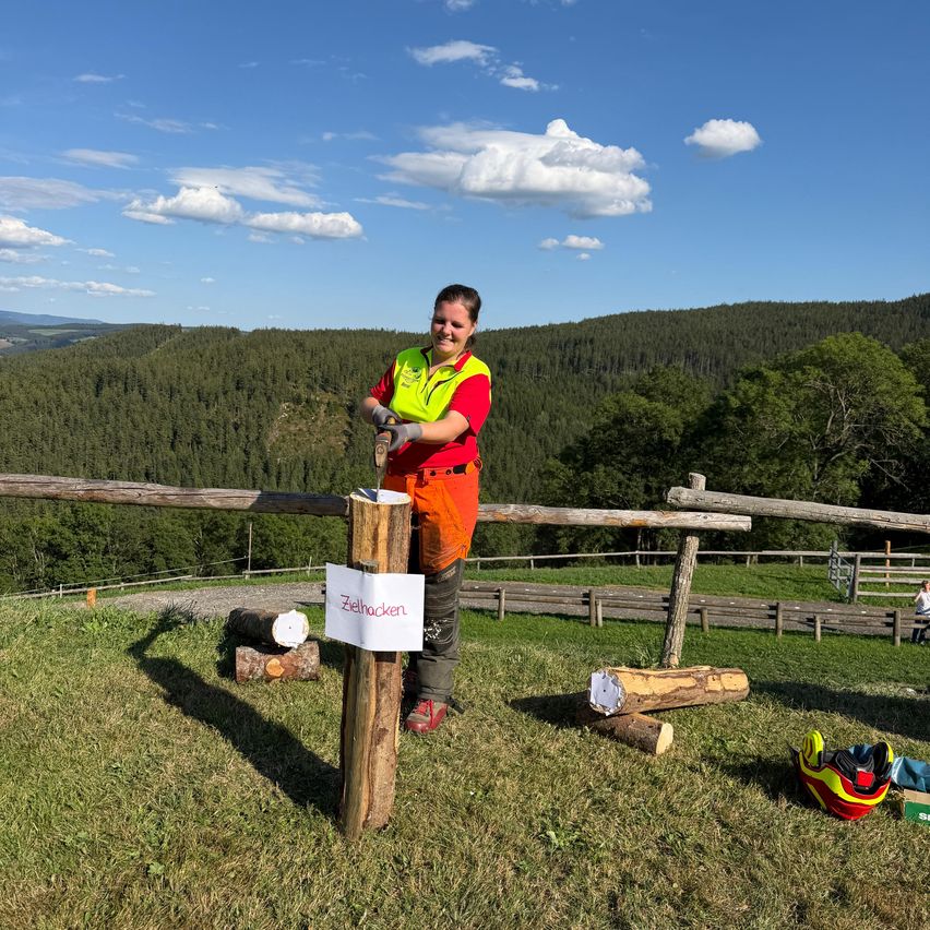 Eine Frau in einer Sicherheitsweste steht neben einem Holzpfosten mit einem Schild, das Zielhacken sagt, umgeben von einem Grasfeld und einer Bergkulisse.