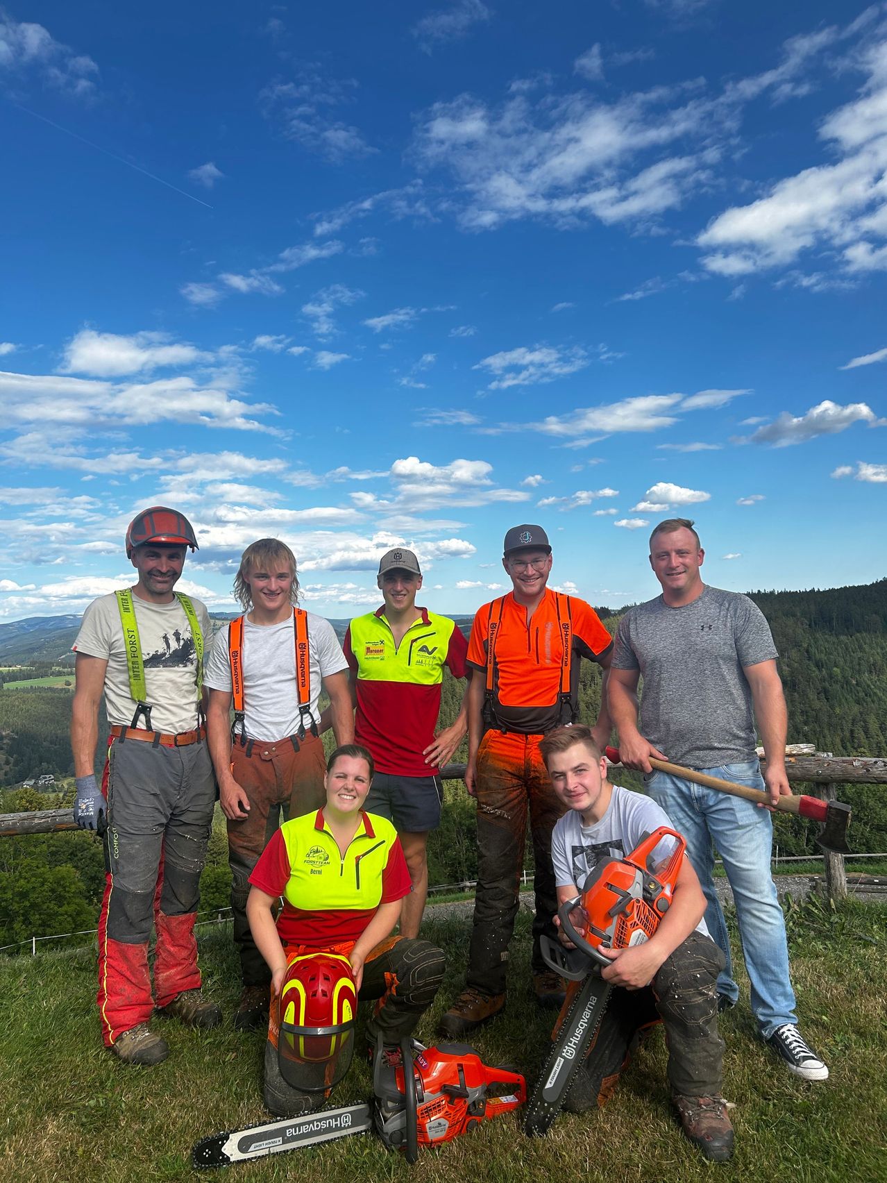 Eine Gruppe von sieben Personen, die in Sicherheitsausrüstung gekleidet sind, stehen auf einem Grashügel mit einem Holzzaun im Hintergrund, unter einem blauen Himmel mit verstreuten Wolken.