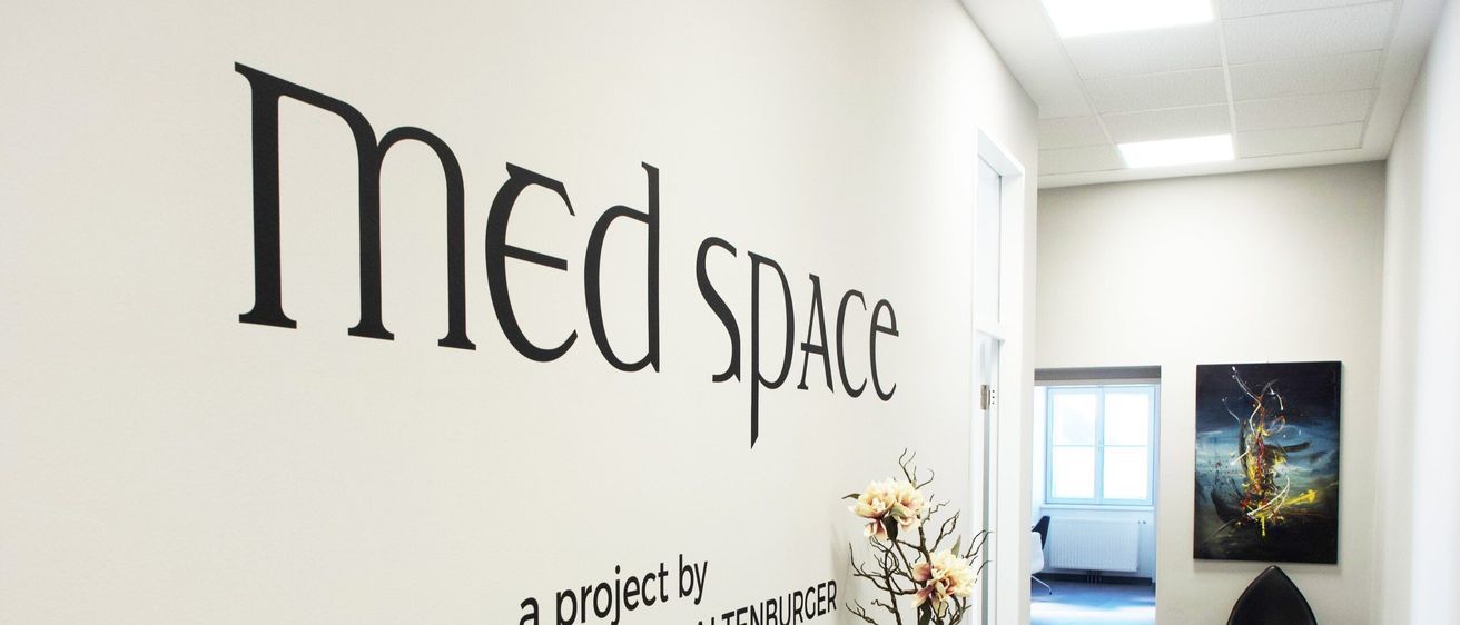 Ein weißer Raum mit einer Wand, die mit 'med space' beschriftet ist. Es gibt einen schwarzen Stuhl, einen Tisch mit Blumen und ein Gemälde an der Wand.