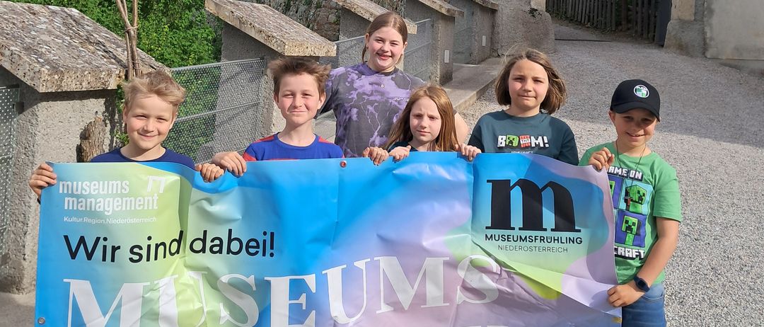 Eine Gruppe von Kindern hält ein Banner mit dem Text 'Museumsfruehling' vor einem historischen Gebäude. Die Kinder lächeln und posieren für ein Foto.