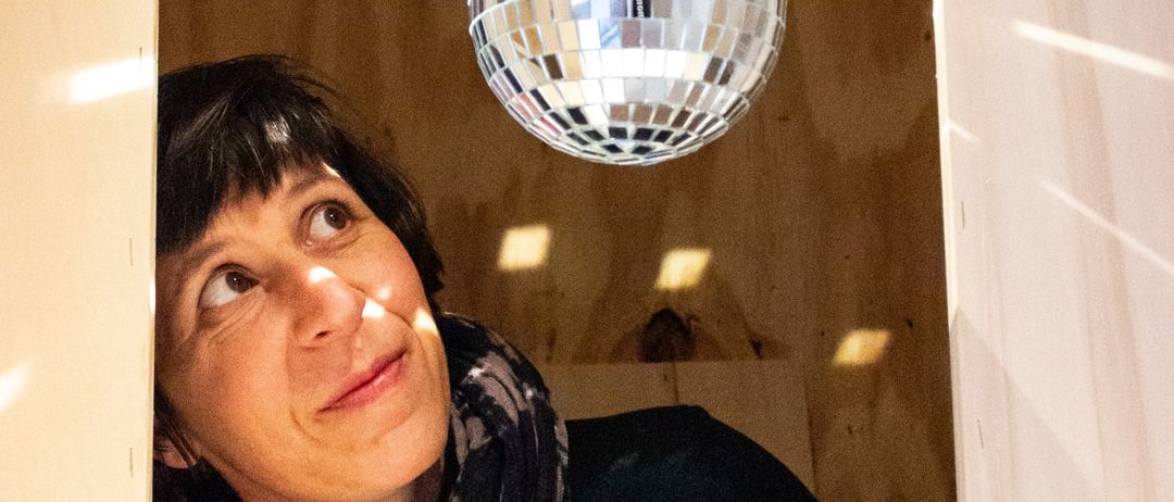 Eine Frau mit einer Schürze lächelt und schaut zu einer Discokugel auf, die von der Decke hängt.