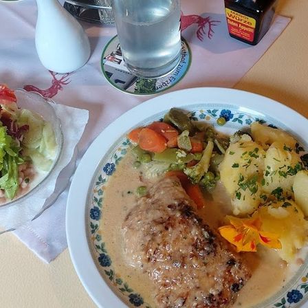 Ein Tisch zum Essen mit einem Teller Schweinekoteletts in cremiger Sauce, begleitet von Gemüse, und einem Glas Wasser.
