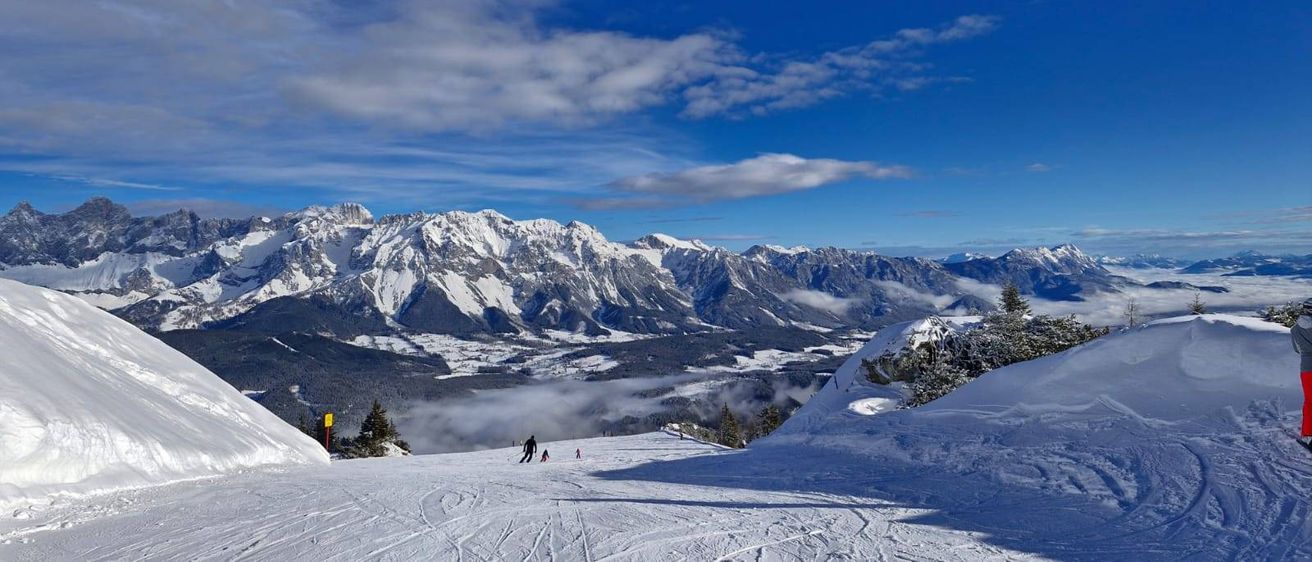 Verschneite Berge mit blauem Himmel. Ein Skifahrer fährt die Piste hinunter. Die Landschaft ist verschneit, mit einigen Bäumen und Skifahrern.