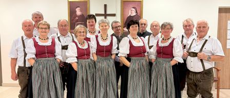 Eine Gruppe von Menschen in traditioneller deutscher Kleidung, die in einer Reihe stehen, mit einem Kreuz und religiösen Gemälden im Hintergrund.