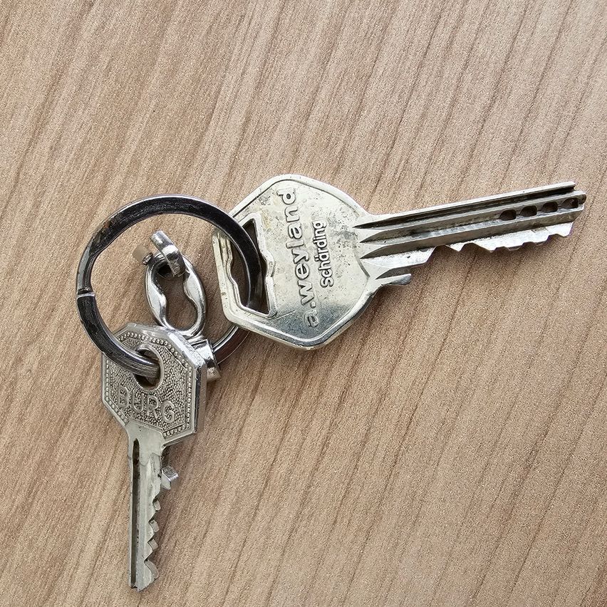 Ein Paar silberner Schlüssel, einer mit der Aufschrift 'Burg' und der andere mit 'Qualawen,' ist an einem Metallring auf einer hölzernen Oberfläche befestigt.