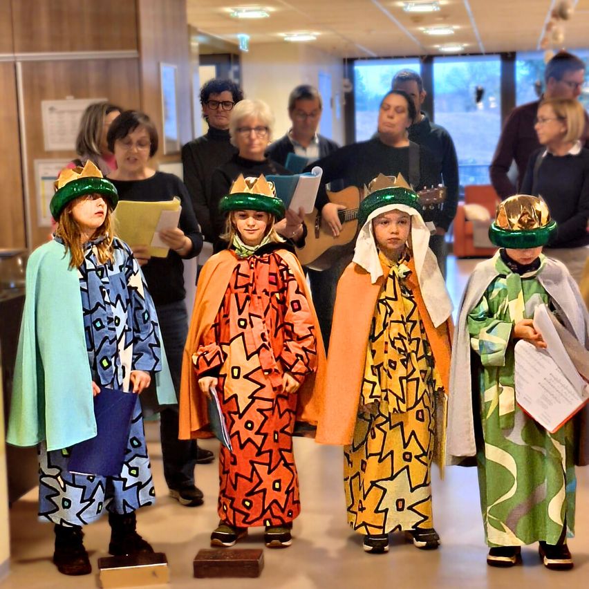 Mehrere Kinder in Kostümen stehen in einem Flur. Vier von ihnen tragen Kronen. Hinter ihnen halten Menschen Papiere und eine Person spielt Gitarre.