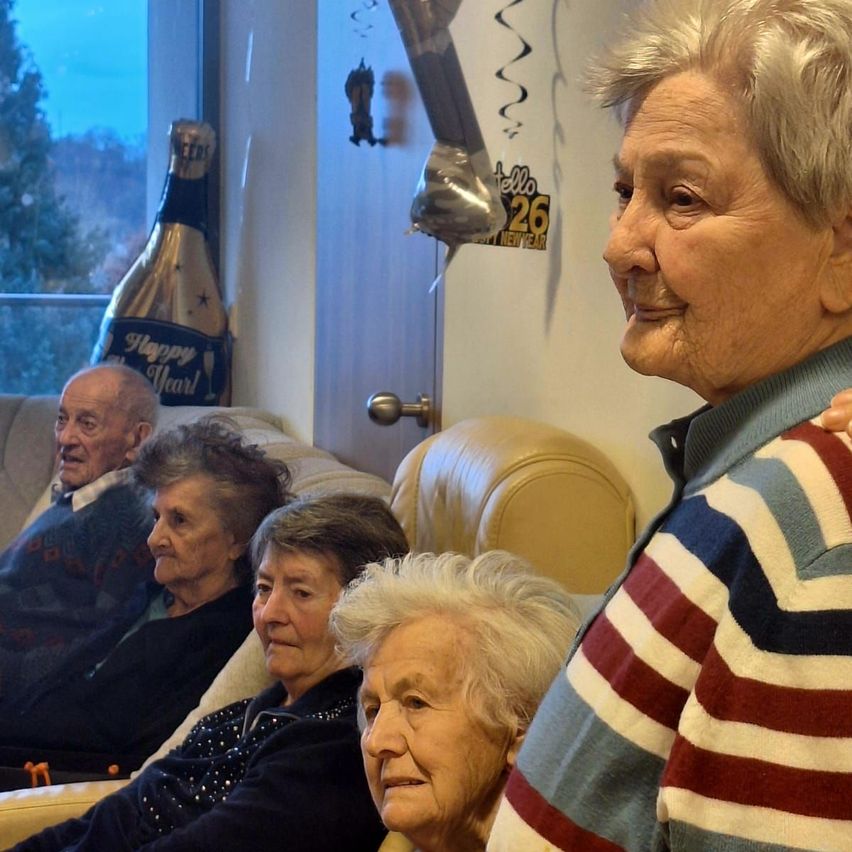 Mehrere ältere Personen sitzen auf einer Couch in einem Wohnzimmer, eine lächelt und schaut in die Kamera. Hinter ihnen ist eine Wand mit einer Champagnerflasche und einem 'Frohes Neues Jahr'-Ballon.