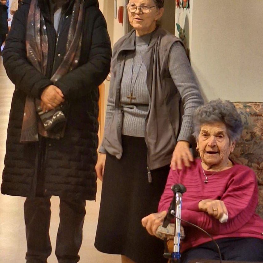 Drei Frauen stehen in einem Flur, eine sitzt auf einem Stuhl. Die Frau auf der linken Seite trägt einen Schal und hält eine Handtasche. Die mittlere Frau trägt eine Brille und eine Weste. Die ältere Frau auf der rechten Seite sitzt mit einem Stock auf einem Stuhl.