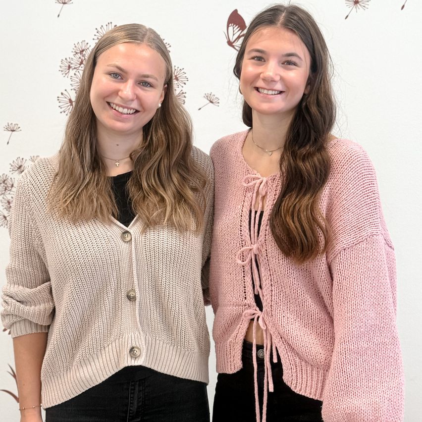 Zwei Frauen stehen eng nebeneinander vor einer weißen Wand mit Blumentapete, beide tragen gestrickte Pullover. Die Frau auf der linken Seite trägt einen beigen Cardigan mit Knöpfen und eine Halskette. Die Frau auf der rechten Seite trägt einen rosa Pullover mit Schnürung. Beide lächeln.