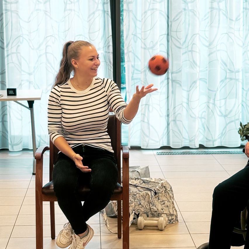 Eine Frau in einem gestreiften Shirt sitzt in einem Stuhl und wirft einen Ball in die Luft. Sie lächelt. Neben ihr sitzt eine andere Person. Ein Schreibtisch und eine Tasche sind im Hintergrund.