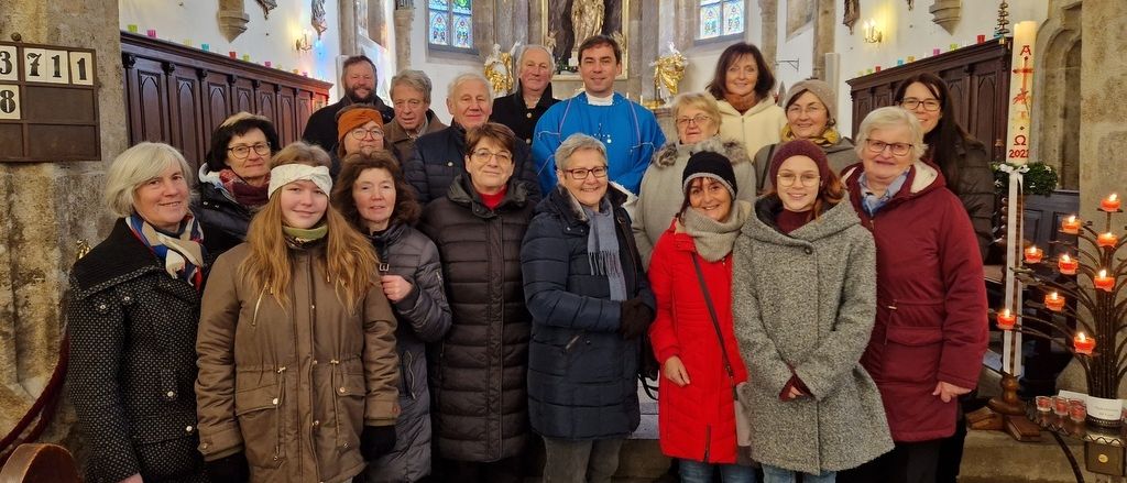 Eine Gruppe von Erwachsenen, die warm gekleidet sind, steht vor einem Kirchenaltar und lächelt für ein Foto. Der Altar hat eine goldene Statue und Buntglasfenster.
