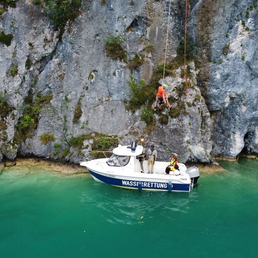 Ein weißes Boot mit blauer Umrandung liegt in einem See. Eine Person mit Helm klettert einen Felsen hinauf. Eine weitere Person steht im Boot und eine dritte Person sitzt im Boot.