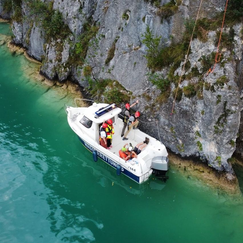 Ein Bergrettungsteam in Helmen bereitet sich darauf vor, eine Person von einem Felsen zu einem Boot auf einem See zu bergen.