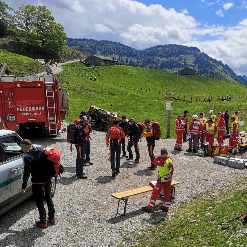 Ein roter Feuerwehrwagen ist am Straßenrand geparkt. Ein umgekipptes Auto ist in der Nähe. Rettungskräfte sind vor Ort, einige tragen Sicherheitswesten und Helme. Eine Holzbank und Tische sind aufgestellt. Ein Berg ist im Hintergrund.