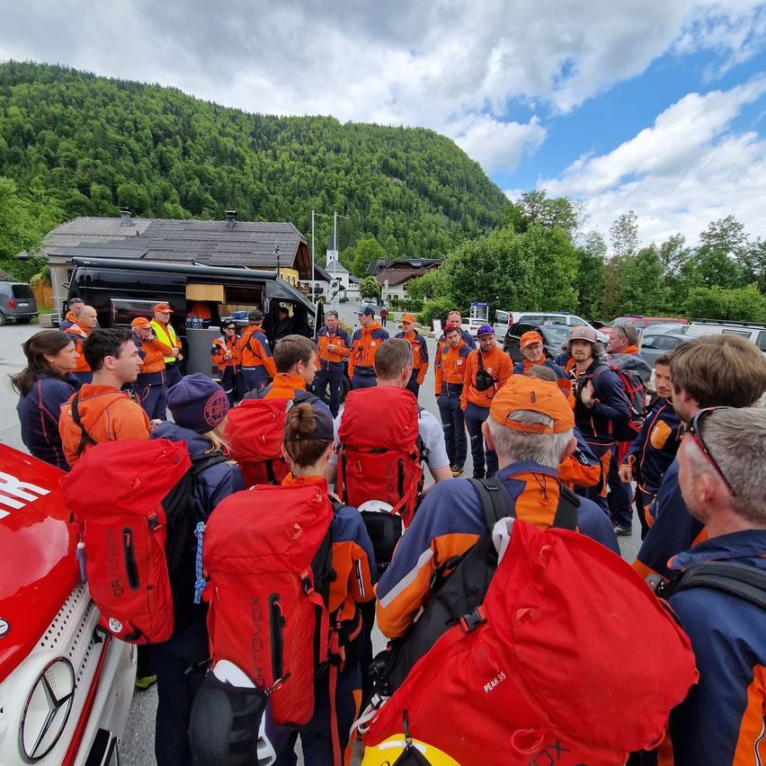 Eine Gruppe von Menschen in orangefarbenen Westen und Mützen versammelt sich im Freien, wahrscheinlich für eine Veranstaltung. Sie stehen um ein Fahrzeug herum und einige tragen rote Rucksäcke. Im Hintergrund befindet sich ein Haus, Autos und ein Berg.