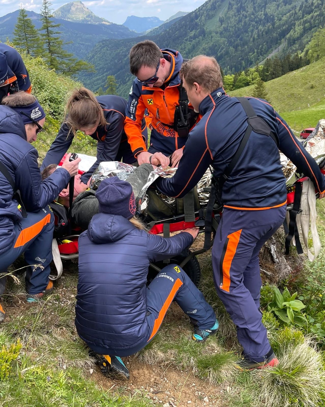 Ein Team von Rettungskräften in blauen und orangefarbenen Uniformen rettet eine verletzte Person aus einem Gebirgsgebiet. Sie benutzen eine Trage und eine Aluminiumdecke.