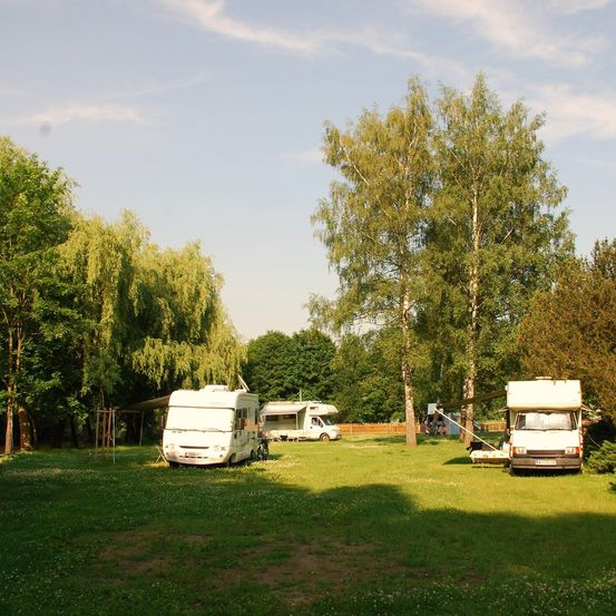 Ein sonniger Tag mit zwei geparkten weißen Wohnmobilen auf einer Rasenfläche, umgeben von Bäumen und einem Holzzaun.