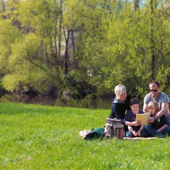 Eine vierköpfige Familie sitzt auf einem Rasenfeld in der Nähe eines Flusses und genießt einen sonnigen Tag. Die Mutter und der Vater lesen ihren Kindern ein Buch vor.