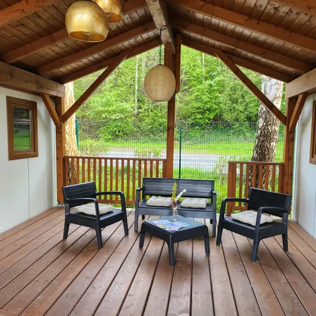 Eine hölzerne Veranda mit gepolsterten Stühlen, einem Tisch und hängenden Laternen. Draußen gibt es einen Blick auf ein Grasfeld mit Bäumen und einem Zaun.